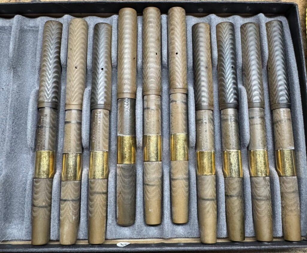 A row of identical vintage hard rubber fountain pens from a shop display, badly faded and discolored to brown from oxidation.